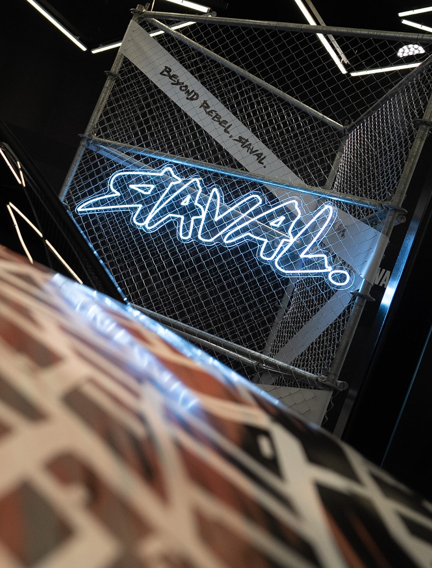 A neon sign spelling "RAVAL!" is displayed on a chain-link fence panel, which also features the text "BEYOND REBEL, RAVAL.". The display is set in a dark, stylized room with geometric lighting patterns.